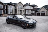 A luxurious black convertible sports car is parked in front of an opulent mansion with large windows and elaborate architectural details. The driveway is expansive, and the overall scene exudes elegance and wealth.