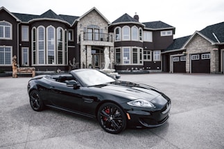 A luxurious black convertible sports car is parked in front of an opulent mansion with large windows and elaborate architectural details. The driveway is expansive, and the overall scene exudes elegance and wealth.