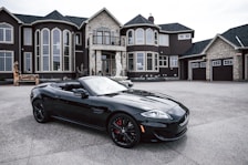 A luxurious black convertible sports car is parked in front of an opulent mansion with large windows and elaborate architectural details. The driveway is expansive, and the overall scene exudes elegance and wealth.