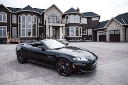 A luxurious black convertible sports car is parked in front of an opulent mansion with large windows and elaborate architectural details. The driveway is expansive, and the overall scene exudes elegance and wealth.