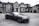 A luxurious black convertible sports car is parked in front of an opulent mansion with large windows and elaborate architectural details. The driveway is expansive, and the overall scene exudes elegance and wealth.