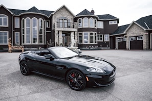 A luxurious black convertible sports car is parked in front of an opulent mansion with large windows and elaborate architectural details. The driveway is expansive, and the overall scene exudes elegance and wealth.