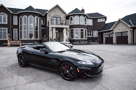 A luxurious black convertible sports car is parked in front of an opulent mansion with large windows and elaborate architectural details. The driveway is expansive, and the overall scene exudes elegance and wealth.