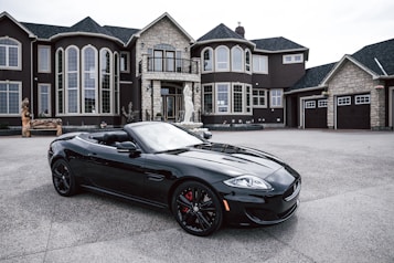 A luxurious black convertible sports car is parked in front of an opulent mansion with large windows and elaborate architectural details. The driveway is expansive, and the overall scene exudes elegance and wealth.