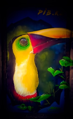A vibrant mural of a toucan with a prominently large yellow and green beak. The bird's eye is large and expressive, surrounded by shades of green. Bright green leaves are visible, suggesting a rainforest setting, and there are dark blue and black areas creating depth and contrast.