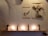 A minimalist wall display features two pieces of artwork hanging on a textured concrete wall. One is an illustration of a squirrel holding a fern, and the other depicts a deer with plant-like antlers. Below, a wooden shelf holds several potted cacti and small candles, casting a warm glow.
