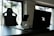 A stylish home office setup with a black gaming chair near a desk. On the desk is a sleek laptop displaying a glowing logo, accompanied by a smartphone resting on a white stand. The background features large windows, allowing natural light to fill the space, highlighting a minimalist and modern aesthetic.