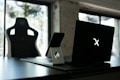 A stylish home office setup with a black gaming chair near a desk. On the desk is a sleek laptop displaying a glowing logo, accompanied by a smartphone resting on a white stand. The background features large windows, allowing natural light to fill the space, highlighting a minimalist and modern aesthetic.