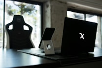 A stylish home office setup with a black gaming chair near a desk. On the desk is a sleek laptop displaying a glowing logo, accompanied by a smartphone resting on a white stand. The background features large windows, allowing natural light to fill the space, highlighting a minimalist and modern aesthetic.