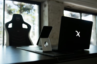 A stylish home office setup with a black gaming chair near a desk. On the desk is a sleek laptop displaying a glowing logo, accompanied by a smartphone resting on a white stand. The background features large windows, allowing natural light to fill the space, highlighting a minimalist and modern aesthetic.