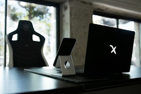 A stylish home office setup with a black gaming chair near a desk. On the desk is a sleek laptop displaying a glowing logo, accompanied by a smartphone resting on a white stand. The background features large windows, allowing natural light to fill the space, highlighting a minimalist and modern aesthetic.