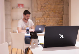 A modern workspace with a focus on technology. In the foreground, there is a black laptop with a distinctive logo placed on a table beside a smartphone propped on a stand. A man is sitting in the background, also using a similar laptop. The setting is minimalistic with a combination of white furniture and a textured brick wall.