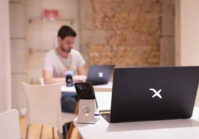 A modern workspace with a focus on technology. In the foreground, there is a black laptop with a distinctive logo placed on a table beside a smartphone propped on a stand. A man is sitting in the background, also using a similar laptop. The setting is minimalistic with a combination of white furniture and a textured brick wall.