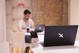 A modern workspace with a focus on technology. In the foreground, there is a black laptop with a distinctive logo placed on a table beside a smartphone propped on a stand. A man is sitting in the background, also using a similar laptop. The setting is minimalistic with a combination of white furniture and a textured brick wall.