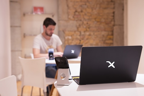 A modern workspace with a focus on technology. In the foreground, there is a black laptop with a distinctive logo placed on a table beside a smartphone propped on a stand. A man is sitting in the background, also using a similar laptop. The setting is minimalistic with a combination of white furniture and a textured brick wall.