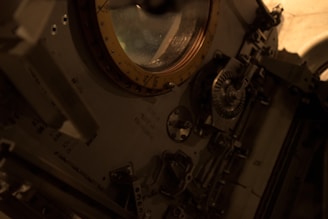 Interior view of a spacecraft module featuring a circular hatch with a golden rim. Surrounding the hatch are various mechanical components, gears, and control panels, giving it a complex and technical appearance.