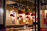 Inside a cozy restaurant with warm lighting, red pendant lamps hang above tables. Wooden shelving units display books and decorative items. A man sits alone at a table, engrossed in his phone, while bowls and eating utensils are set out on the tables.