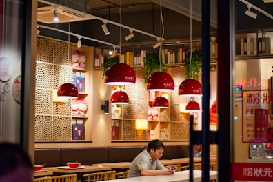 Inside a cozy restaurant with warm lighting, red pendant lamps hang above tables. Wooden shelving units display books and decorative items. A man sits alone at a table, engrossed in his phone, while bowls and eating utensils are set out on the tables.