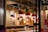 Inside a cozy restaurant with warm lighting, red pendant lamps hang above tables. Wooden shelving units display books and decorative items. A man sits alone at a table, engrossed in his phone, while bowls and eating utensils are set out on the tables.