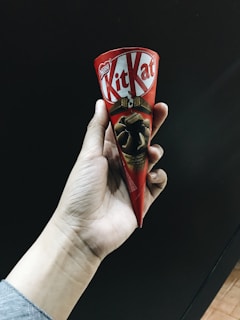 A hand holding a Kit Kat chocolate cone with a red wrapper. The product features a design showing pieces of chocolate on it.