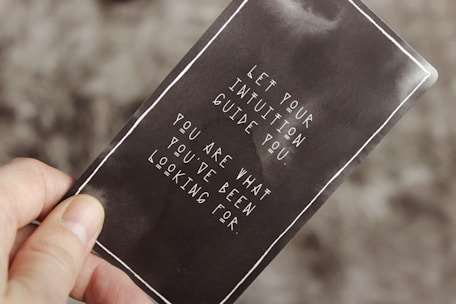 A hand holding a card with a motivational quote in white text against a dark background. The quote reads: 'Let your intuition guide you. You are what you've been looking for.'