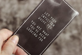 A hand holding a card with a motivational quote in white text against a dark background. The quote reads: 'Let your intuition guide you. You are what you've been looking for.'