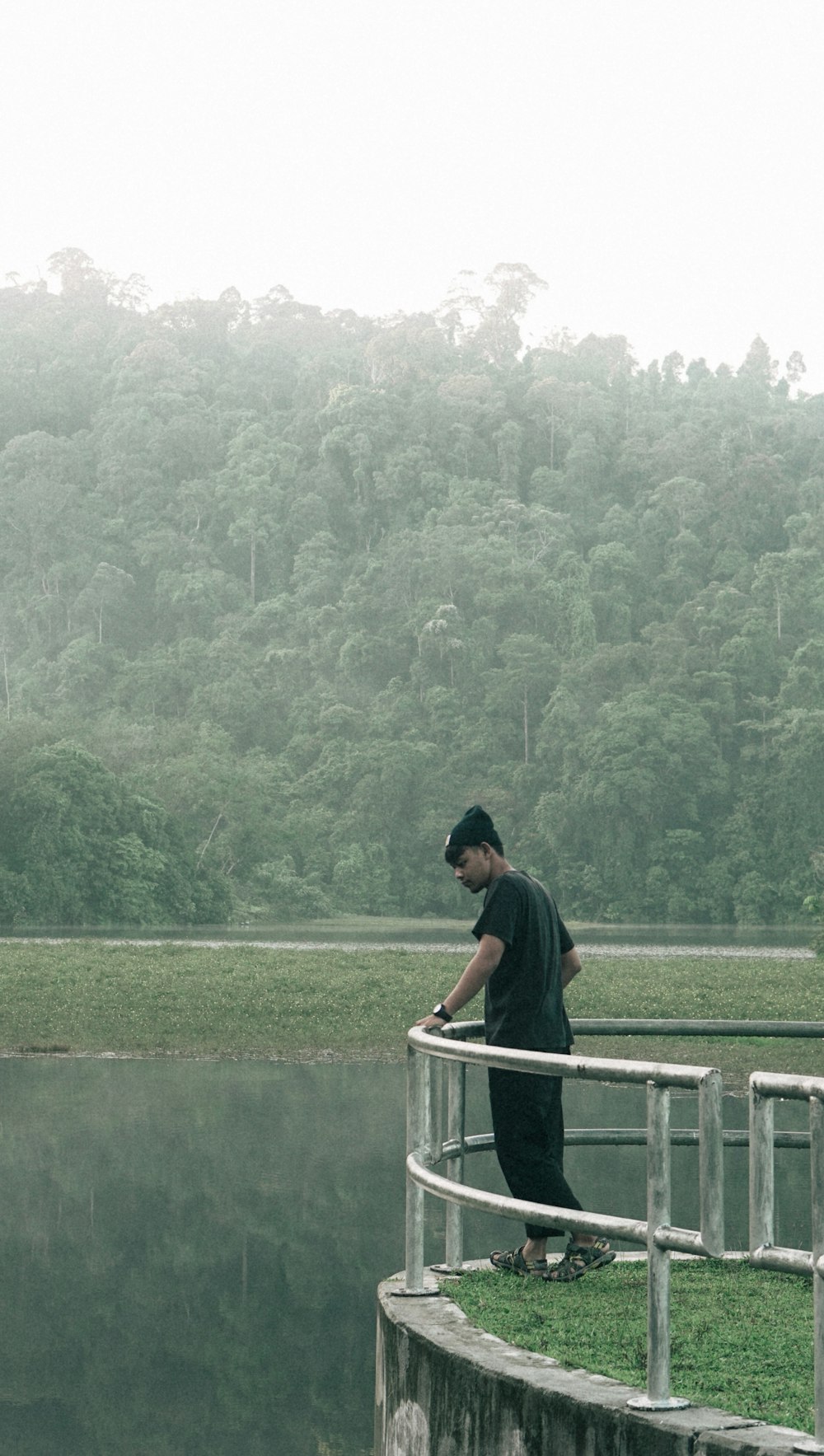 man wearing black crew-neck t-shirt and black hat standing and holding on gray stainless steel railings looking down on body of water surrounded with tall and green trees during daytime