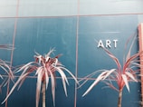 A minimalist composition featuring reddish-brown tropical plants against a deep teal tiled wall. The letters 'ART' are prominently displayed on the right side of the wall.