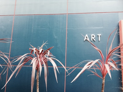 A minimalist composition featuring reddish-brown tropical plants against a deep teal tiled wall. The letters 'ART' are prominently displayed on the right side of the wall.