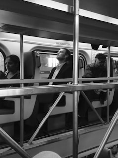 Several passengers are seated inside a train, with a metal railing structure in the foreground. The passengers appear relaxed, with one person resting with their eyes closed. The interior of the train is visible, including signs, seats, and windows.