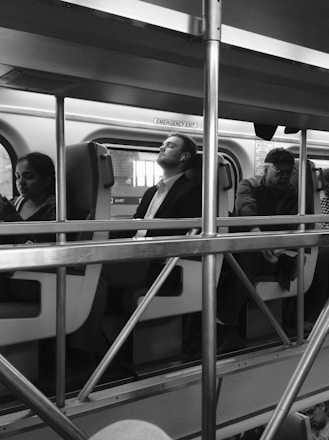 Several passengers are seated inside a train, with a metal railing structure in the foreground. The passengers appear relaxed, with one person resting with their eyes closed. The interior of the train is visible, including signs, seats, and windows.