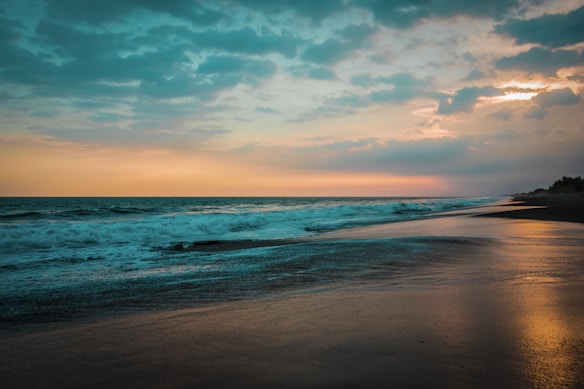 The image depicts a serene beach at sunset with gentle waves lapping against the shore. The sky is painted with warm hues of orange and pink, contrasting against the darker tones of the incoming night. Fluffy clouds partially cover the sun, creating a dramatic play of light and shadow on the sand.
