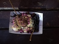 A paper tray holds a dessert consisting of balls of dough topped with chocolate sauce, crushed nuts, and colorful sprinkles. Two wooden sticks are inserted into the dessert. The tray is placed on a dark wooden surface.
