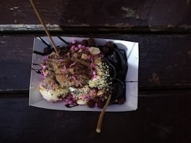 A paper tray holds a dessert consisting of balls of dough topped with chocolate sauce, crushed nuts, and colorful sprinkles. Two wooden sticks are inserted into the dessert. The tray is placed on a dark wooden surface.