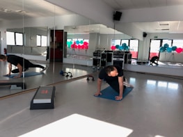 A person is exercising on a mat in a bright room with mirrors on the wall. Various fitness equipment such as weights and kettlebells are scattered on the floor. There are colorful exercise balls stacked in the corner near the windows. The room is well-lit by natural light coming through the windows.
