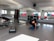 A person is exercising on a mat in a bright room with mirrors on the wall. Various fitness equipment such as weights and kettlebells are scattered on the floor. There are colorful exercise balls stacked in the corner near the windows. The room is well-lit by natural light coming through the windows.