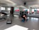 A person is exercising on a mat in a bright room with mirrors on the wall. Various fitness equipment such as weights and kettlebells are scattered on the floor. There are colorful exercise balls stacked in the corner near the windows. The room is well-lit by natural light coming through the windows.