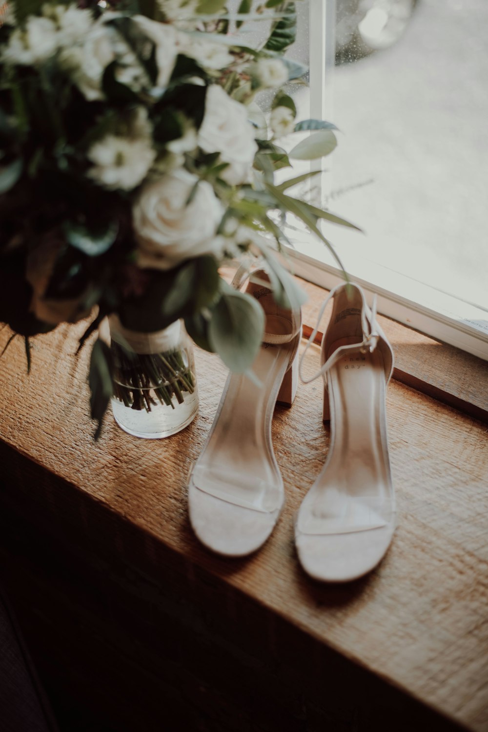 tacones blancos con tira abierta al tobillo junto al jarrón de flores