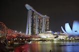 A night view of a vibrant urban landscape featuring a modern, iconic building complex with three towers supporting a rooftop platform shaped like a ship. To the right, a uniquely designed structure with a petal-like appearance is illuminated. In the foreground, a futuristic bridge with a spiraling design is lit in red, spanning across a body of water reflecting the city lights.