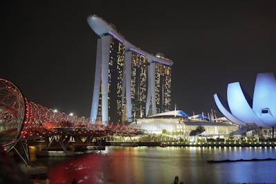 A night view of a vibrant urban landscape featuring a modern, iconic building complex with three towers supporting a rooftop platform shaped like a ship. To the right, a uniquely designed structure with a petal-like appearance is illuminated. In the foreground, a futuristic bridge with a spiraling design is lit in red, spanning across a body of water reflecting the city lights.