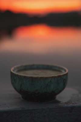 A ceramic bowl with a textured teal glaze sits in the foreground, capturing a soft, warm glow from an out-of-focus sunset in the background. The sky displays vibrant orange and pink hues, fading into darker shades.