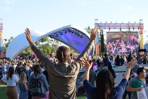 A large outdoor event with a crowd of people gathered in front of a stage. The stage is covered by a white canopy, and bright lights illuminate it. Attendees are raising their hands, suggesting enthusiasm or participation. A large screen displays an image of another part of the event, contributing to the lively atmosphere. The surrounding area is a grassy park-like setting with trees in the background.