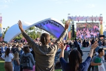 A large outdoor event with a crowd of people gathered in front of a stage. The stage is covered by a white canopy, and bright lights illuminate it. Attendees are raising their hands, suggesting enthusiasm or participation. A large screen displays an image of another part of the event, contributing to the lively atmosphere. The surrounding area is a grassy park-like setting with trees in the background.