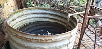 A large, rusting metal barrel filled with water and some floating debris is placed outdoors, surrounded by lush vegetation. A pipe extends into the barrel, likely used for filling it with water. The area around the barrel has an unkempt look with metal and wooden structures nearby.