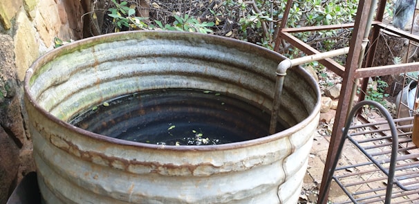 A large, rusting metal barrel filled with water and some floating debris is placed outdoors, surrounded by lush vegetation. A pipe extends into the barrel, likely used for filling it with water. The area around the barrel has an unkempt look with metal and wooden structures nearby.