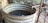 A large, rusting metal barrel filled with water and some floating debris is placed outdoors, surrounded by lush vegetation. A pipe extends into the barrel, likely used for filling it with water. The area around the barrel has an unkempt look with metal and wooden structures nearby.