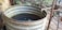 A large, rusting metal barrel filled with water and some floating debris is placed outdoors, surrounded by lush vegetation. A pipe extends into the barrel, likely used for filling it with water. The area around the barrel has an unkempt look with metal and wooden structures nearby.