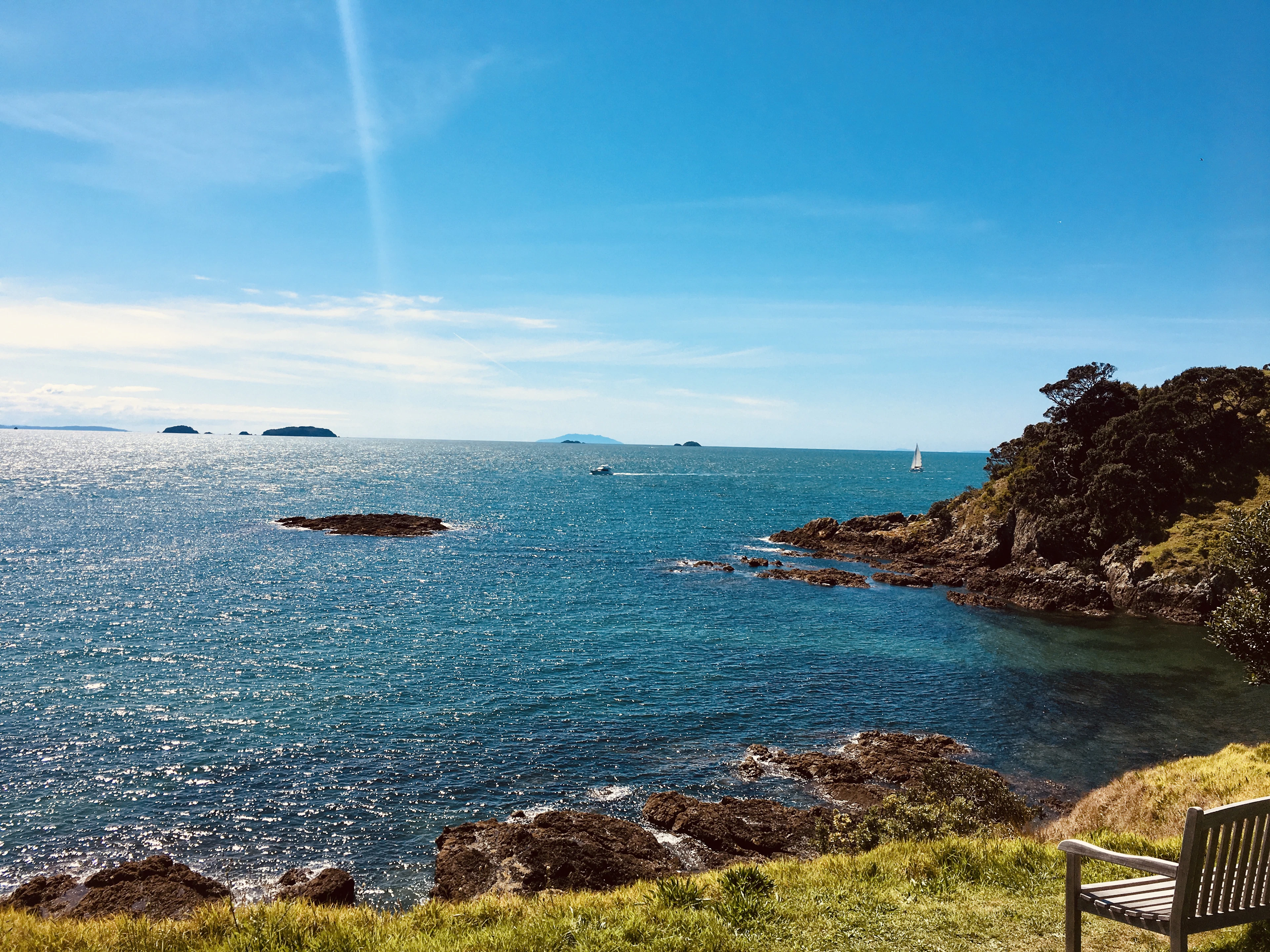 A wooden bench overlooks a rocky Waiheke Island cove with clear blue water and a distant sailboat.