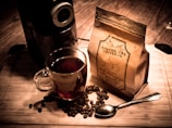 A wooden surface holds a glass cup filled with coffee, surrounded by scattered coffee beans and a shiny silver spoon. A bag labeled 'Apothecary Coffee Co.' stands beside an electric coffee grinder, creating a warm, inviting scene.