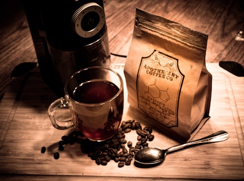 A wooden surface holds a glass cup filled with coffee, surrounded by scattered coffee beans and a shiny silver spoon. A bag labeled 'Apothecary Coffee Co.' stands beside an electric coffee grinder, creating a warm, inviting scene.